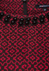 Tissu noir avec un motif floral rouge, ornée d'une rangée de perles noires le long du bord de l'encolure, complétée par une fermeture éclair centrale.