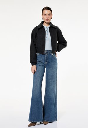 Kvinde iført en sort cropped jakke, lyseblå ribbet polo-shirt, højtragtede vidde jeans og brune spidse sko.