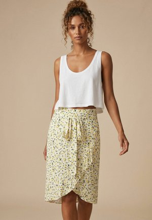 Femme debout, portant un crop top blanc sans manches et une jupe portefeuille jaune à fleurs nouée à la taille, sur un fond beige uni.