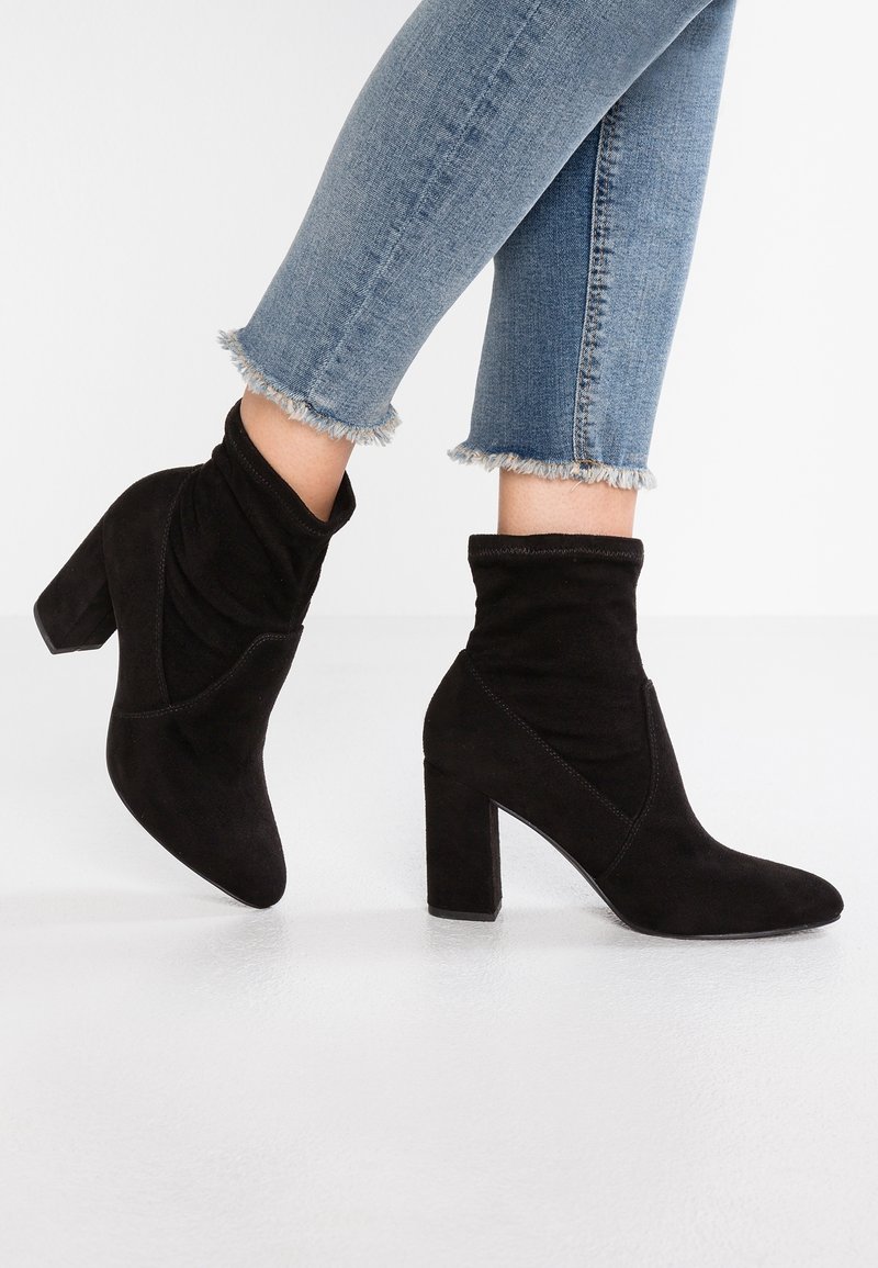 Jambes portant un jean bleu effiloché et des bottines noires en daim à talons épais, posées sur un fond blanc.