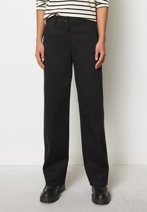 Persoon draagt zwarte hoge rechte broek, oversized zwarte schoenen en een wit overhemd met lange mouwen en zwarte horizontale strepen.