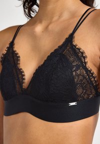 Schwarzer Spitzen-Bralette mit dreieckiger Form, zarten Fransen-Details, dünnen Trägern und einem festen schwarzen Unterband mit kleinem Logotag.