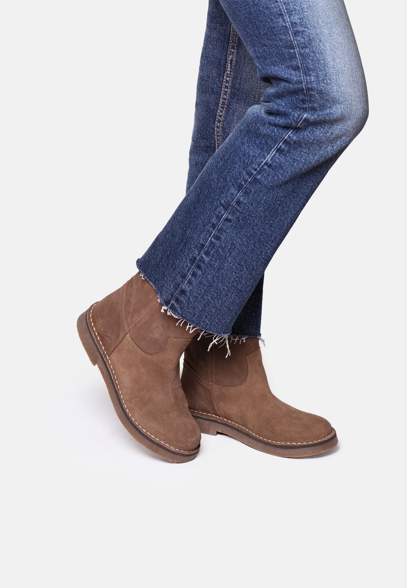 Bottines en cuir suédé marron avec un bout arrondi et des coutures latérales, portées avec un jean en denim à ourlet effiloché dans un cadre décontracté.
