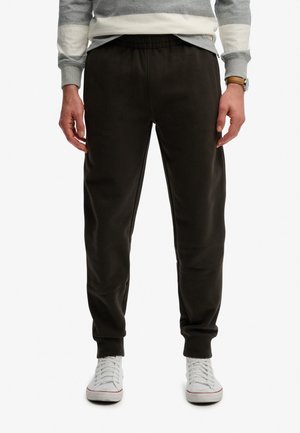 Mann trägt schwarze Jogginghosen, weiße Sneakers, grauen Pullover und eine braune Armbanduhr, steht vor weißem Hintergrund.