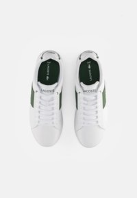 Ett par vita Lacoste-sneakers med gröna sidopaneler och vita skosnören, sedd ovanifrån på en vit bakgrund.