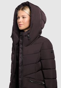 Taukainājs tumši purpursarkanā krāsā ar lielu pildītu kapuci, ar rāvējslēdzēju priekšā un sānu kabatām. Iezīmēts ar quilted dizainu un melnu metāla aksesuāru.
