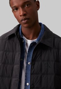 Jachetă neagră matlasată cu guler, nasturi tip snap și un logo subtil "CK", purtată peste o cămașă din denim albastru cu accente argintii.