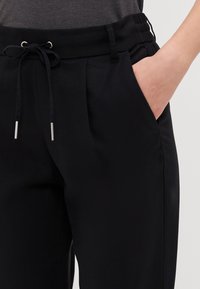 Un pantalon noir avec un cordon de serrage à la taille, des poches latérales et une texture lisse agrémentée de plis pour un détail de design supplémentaire.