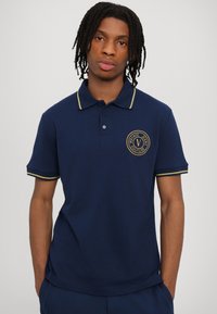 Marineblå polo skjorte laget av bomull, med krage med gul kant og brodert logo på brystet. Korta ermer med matchende kant.