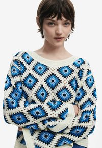 Pull en crochet présentant un motif en diamant dans des tons de bleu, noir et crème. Coupe ample avec un large décolleté et des manches longues. Fini texturé.