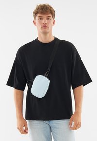 Piccolo borsello a tracolla blu realizzato in materiale liscio, di forma rettangolare, con chiusura a zip e tracolla regolabile, indossato sopra una maglietta nera.