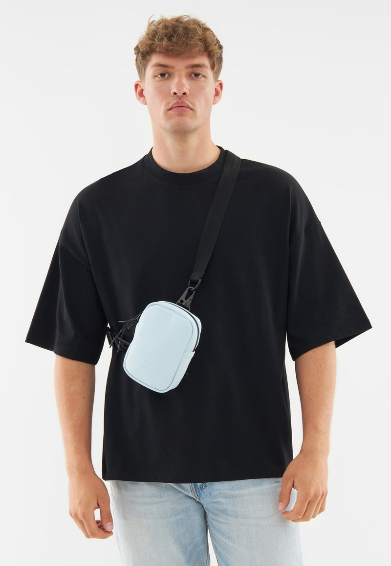 Piccolo borsello a tracolla blu realizzato in materiale liscio, di forma rettangolare, con chiusura a zip e tracolla regolabile, indossato sopra una maglietta nera.