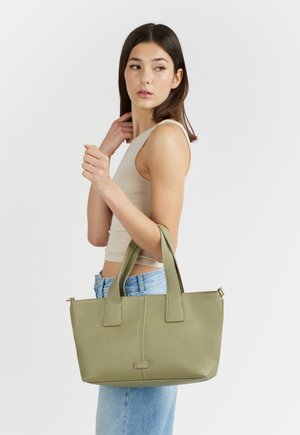 Giovane donna con top beige senza maniche e jeans porta una borsa tote in pelle verde oliva sul braccio, guardando di lato.