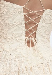 Πίσω όψη ενός κρεμ δαντελένιου φορέματος με λουλουδάτα μοτίβα και δεσίματα με λουριά που σχηματίζουν σταυρό, αποκαλύπτοντας το δέρμα στην πάνω πλάτη.