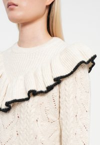 Cream knit sweater with textured pattern, featuring a ruffled collar edged in black. The fabric appears soft and warm.