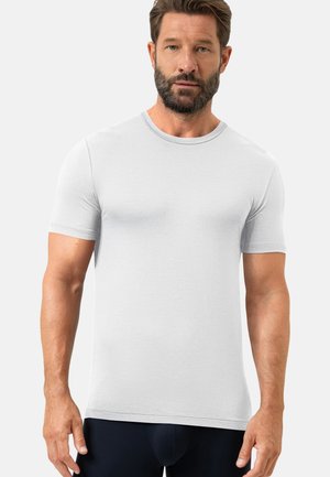 Mężczyzna z brodą, ubrany w jednolity biały t-shirt z krótkim rękawem i ciemne spodnie, stojący na tle jednolitego jasnego tła.
