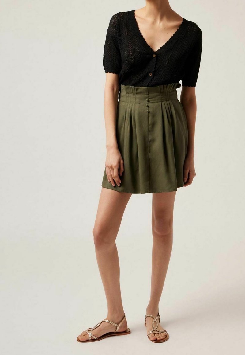 Femme portant un haut en maille noir à manches courtes avec des boutons, une jupe plissée taille haute vert olive, et des sandales plates beiges.