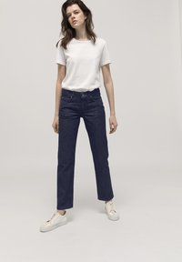 Camiseta de algodón blanca, jeans oscuros de corte alto y recto, combinados con zapatillas de color claro sobre un fondo neutro.