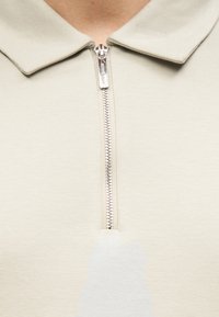 Ανοιχτό γκρι πόλο με μισό φερμουάρ και απαλό γιακά. Το ύφασμα φαίνεται λείο με διακριτική γυαλάδα· διαθέτει ξεθωριασμένο γραφικό σχέδιο στο μπροστινό μέρος.