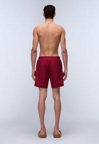 Shorts de bain rouges avec une taille élastique, arrivant à mi-cuisse. Le tissu semble léger et lisse, avec une poche arrière.