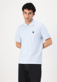 Ανοιχτό μπλε πόλο μπλουζάκι με γιακά, κοντά μανίκια και ένα μικρό μαύρο έμβλημα στο αριστερό στήθος. Συνδυασμένο με σκούρες τζιν.