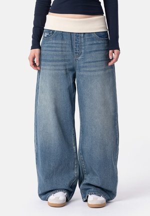 Person trägt weite blaue Jeans mit weißem elastischem Bund und weißen Sneakers und steht vor einem schlichten Hintergrund.
