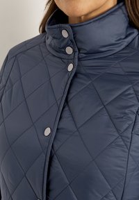 Marineblau gesteppte Jacke mit hohem Kragen, ausgestattet mit silbernen Druckknöpfen und einer glatten, glänzenden Textur.