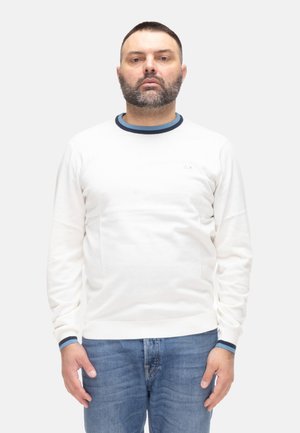 Uomo con barba corta che indossa una camicia bianca a maniche lunghe con rifiniture blu e jeans blu, in piedi dritto contro uno sfondo chiaro uniforme.