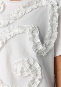 Vit t-shirt med texturerad tyg och dekorativa vita volanger i spiralformade mönster på framsidan. Korta ärmar och rund halsringning.