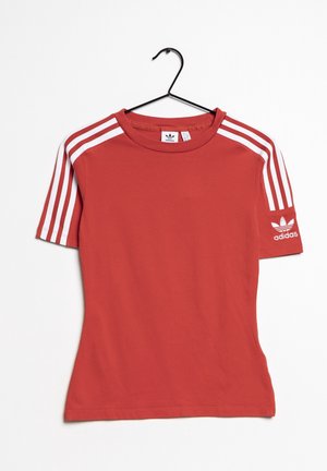 Červené bavlněné tričko s bílým designem tří pruhů na ramenou a vyšitým logem na rukávu. Krátké rukávy a kulatý výstřih.