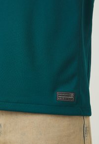 Teal, textured athletic shirt with a tag displaying "ENGINEERED" and numbers, paired with light beige fabric below.