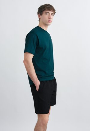 Ung mann med krøllete hår iført mørkegrønn kortermet skjorte og svarte shorts, stående med hendene i lommene mot en hvit bakgrunn.