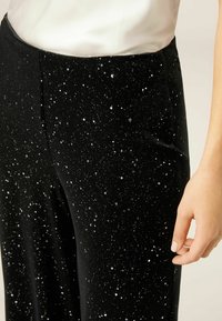 Zwarte fluwelen broek met een verspreid sterrenpatroon en zilveren spikkels. De stof heeft een zachte textuur en het ontwerp heeft een wijde pijp.