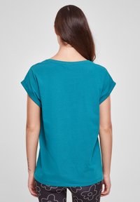 Teal majica s kratkimi rokavi z okroglim ovratnikom, sproščenim krojem in zavitimi rokavi. Mehka bombažna tkanina brez vidnih vzorcev ali okraskov.