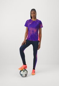 Camisola desportiva respirável roxa com detalhes em rosa, combinada com leggings azul-marinho com painéis laterais roxos e sapatilhas de futebol cor-de-rosa brilhante.
