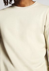 Jasnobeżowa bluza z okrągłym dekoltem, długimi rękawami i gładką fakturą. Brak widocznych wzorów i detali metalowych.