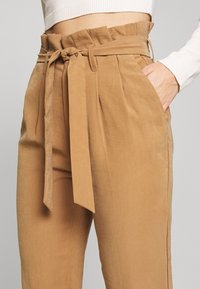 Pantalon marron clair, taille haute, avec ceinture et design plissé. Texture lisse et poches latérales visibles.