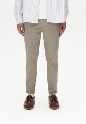 Mężczyzna ubrany w beżowe spodnie z podwiniętymi nogawkami, białe skarpetki, brązowe skórzane buty oraz pasiastą koszulę z długim rękawem, stojący na białym tle.