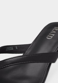 Sandale en cuir noir avec fines lanières et semelle cousue, montrant une partie du nom de la marque "RAID" sur la semelle intérieure.