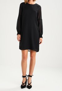 Schwarzes Kleid mit transparenten langen Ärmeln, runder Ausschnitt und geradem Schnitt. Verfügt über geraffte Manschetten und einfarbige Gestaltung. Kombiniert mit schwarzen Riemchen-Heels.