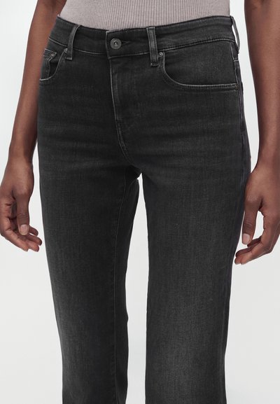 Μαύρα τζιν από denim που φοράει ένα άτομο με τα χέρια χαλαρά στο πλάι, μπροστά από ένα απλό φόντο.