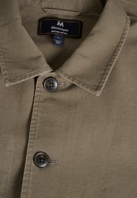 Ruda marškinių su plačiu apykakle, su mygtukų uždarymu ir tamsiu mygtuku. Etiketėje parašyta "Matinique Heritage Apparel" kaip prekės ženklo identifikacija.