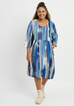Blue-patterned dress with three-quarter sleeves, slight waist seam, pockets, and a loose fit. Worn with beige lace-up boots.