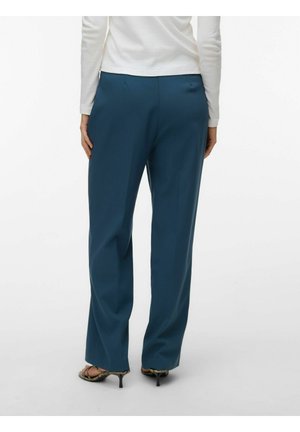 Frau in locker sitzender, hoch taillierter türkiser Hose und weißem Langarmoberteil, steht mit entspannten Händen an den Seiten, Rücken zum Betrachter.