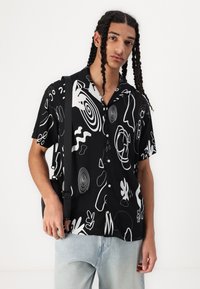 Svart kortermet skjorte med et hvitt abstrakt mønster, knappefeste, lett stoff og avslappet passform, kombinert med lyseblå jeans.