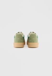 Grønne ruskindsneakers med en polstret krave og en beige gummisål. Mærket "ARKK" er præget på hælen.
