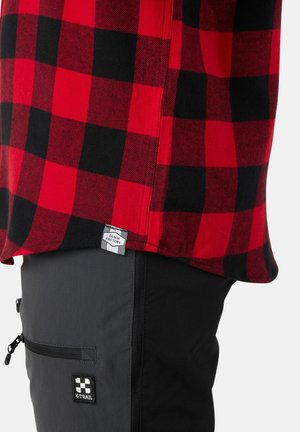 Rood en zwart geblokt shirtstof met een label; gecombineerd met grijze broek met zakken en een logo-patch op de dij.