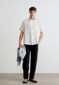 Νέος άνδρας που φοράει λευκό πουκάμισο με κοντά μανίκια πάνω από λευκό μπλουζάκι, μαύρο παντελόνι, μπεζ παπούτσια, κρατώντας ανοιχτό γαλάζιο μπουφάν στο αριστερό χέρι, όρθιος.