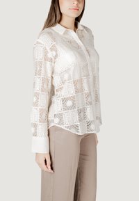 Läbipaistvast valgest kangast, ruudulise mustriga pitspluus, millel on kaelus ja pikad varrukad. Pluusi esituses on kindel valge mansett.