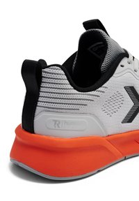 Grijze sportschoen met een gestructureerd bovenwerk, zwarte accenten en een oranje rubberen zool. Heeft een logo aan de zijkant en een trekstropje aan de hiel.